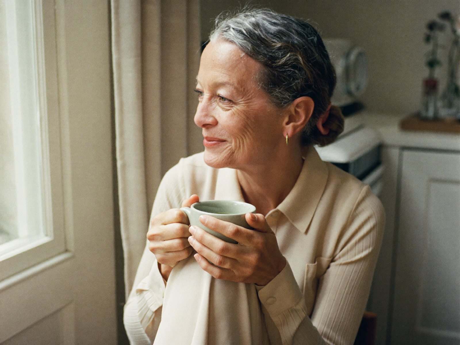 A woman in her late forties sits by a sunlit window with a warm mug of tea, a calm early-morning moment.