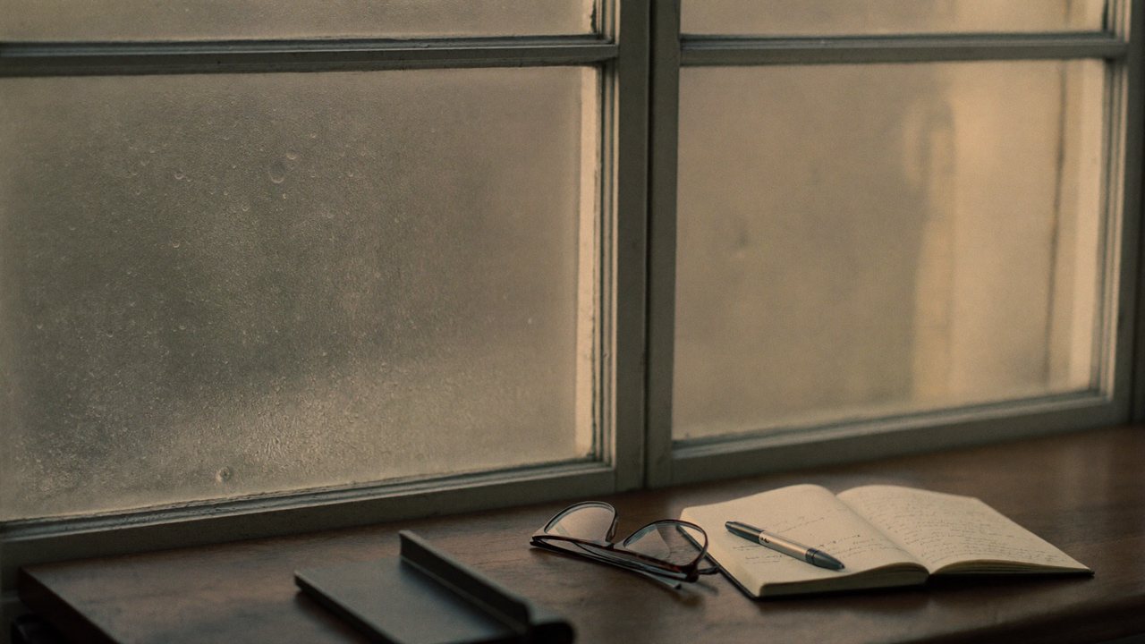 A window clouded with morning condensation beside an open notebook, pen, and reading glasses.
